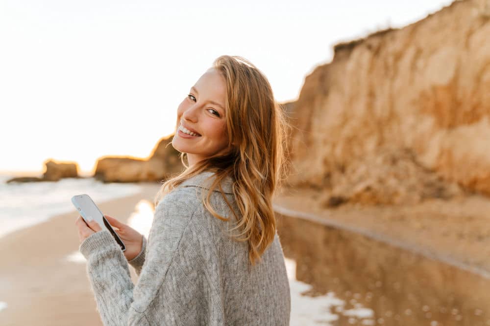 a-woman-smiling-on-the-beach-holding-her-phone